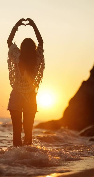 Femme devant un couché de soleil, les pieds dans la mer et faisant un coeur avec les mains.